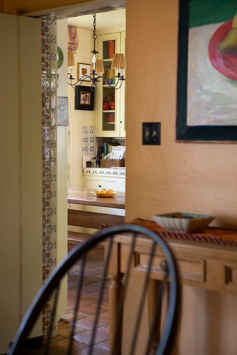 color interior photo, looking through a doorway, chair in foreground, orange walls, tiled floor, a hanging lamp over a table with fruit on it
